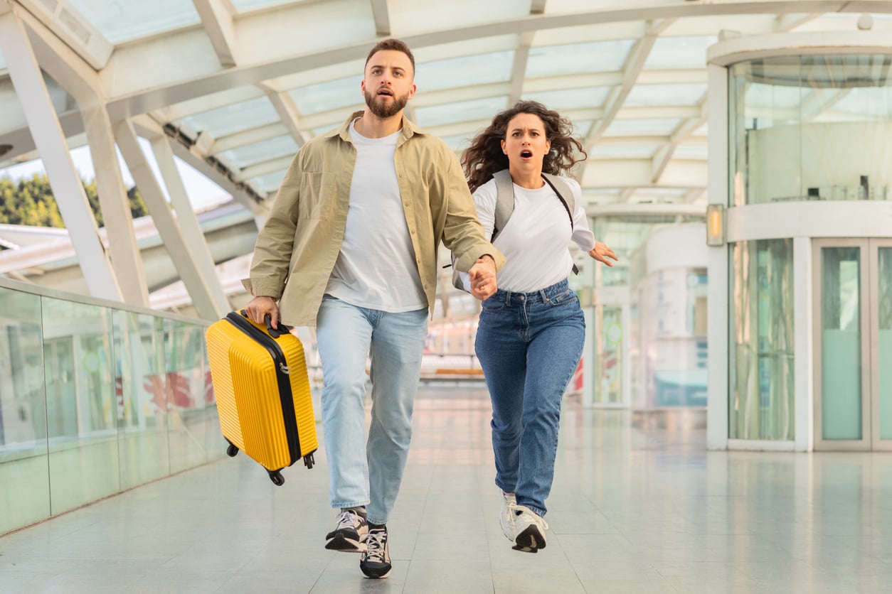 A man and woman holding hands run through a modern glass walkway. The man carries a yellow suitcase, and both look surprised or worried. They are wearing casual clothes and appear to be in a rush.