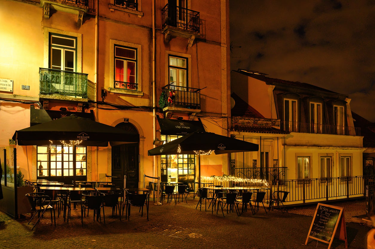 Outdoor tables and chairs sit under black umbrellas at a warmly lit café on a quiet street at night, with string lights decorating one table and two buildings in view—a hidden gem where travelers can relax and avoid common vacation mistakes while soaking in the cozy ambiance.