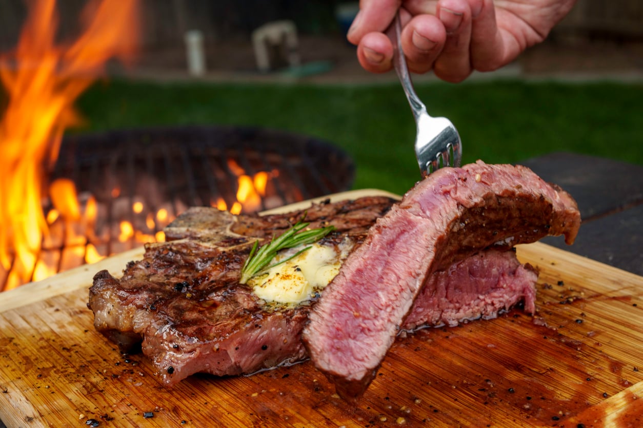 A hand uses a fork to lift a juicy, medium-rare steak with herb butter and rosemary—one of the most mouthwatering foods—on a wooden board near a flaming grill outdoors.