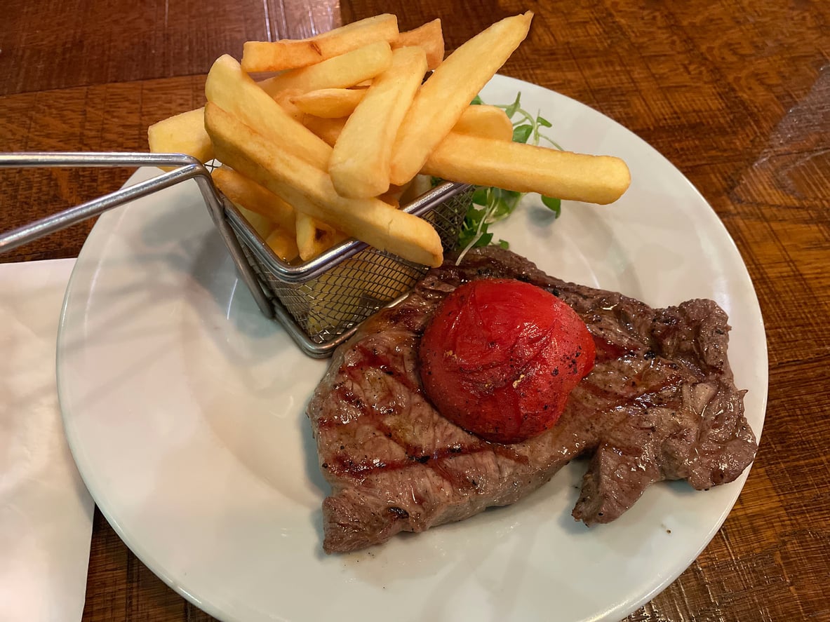 A plate with grilled steak topped with a roasted tomato and a small basket of French fries on the side, served on a wooden table.