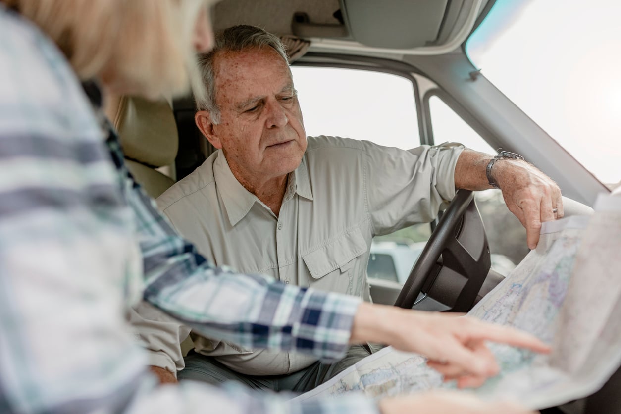 An older man sitting in a vehicle looks closely at a paper map, while a woman beside him points to a location on the map.