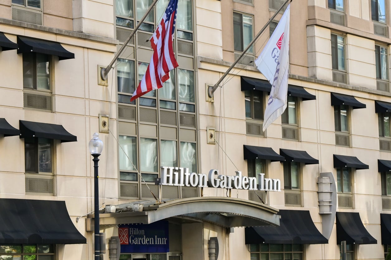 The exterior of a Hilton Garden Inn hotel, part of one of the world’s renowned hotel chains, features the hotel sign above the entrance, two flags, black window awnings, and beige walls.