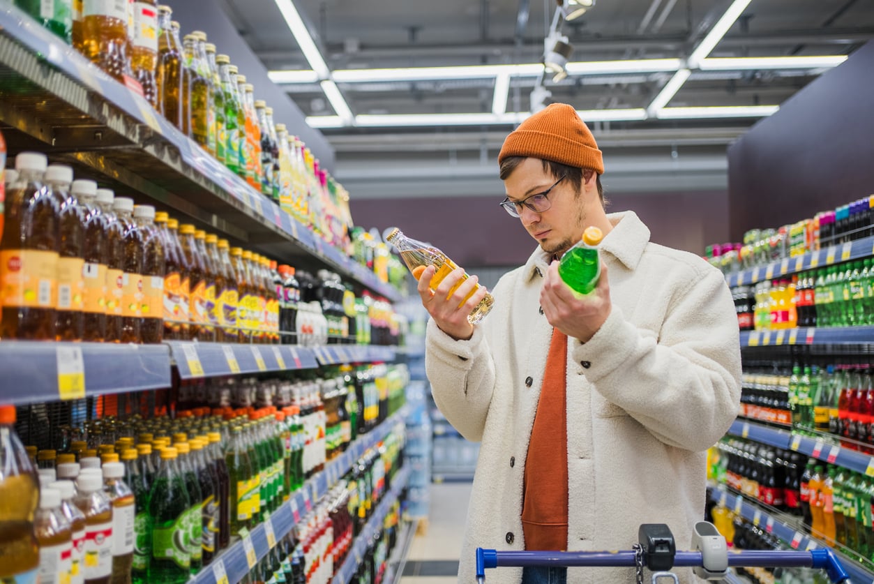 A person in a supermarket aisle holds bottles of beverages, comparing labels as part of their grocery habits, surrounded by shelves full of colorful drink bottles.