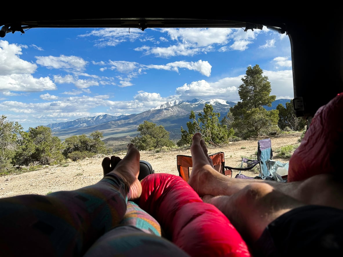 Two people laying in a car with sleeping bags, looking out at a scenic mountain landscape with trees and clouds, camping chairs outside.