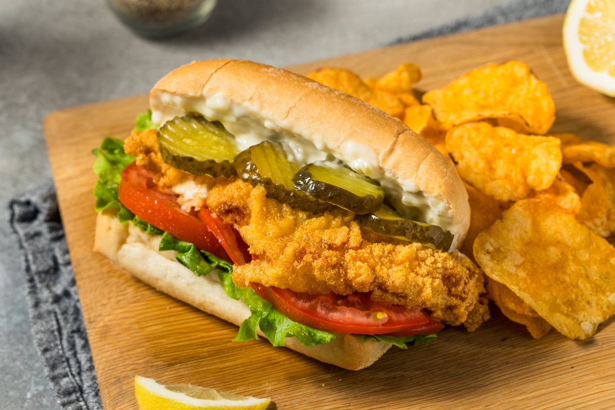 A crispy fried chicken sandwich with pickles, lettuce, tomato, and mayonnaise on a sub roll, served with potato chips on a wooden board.