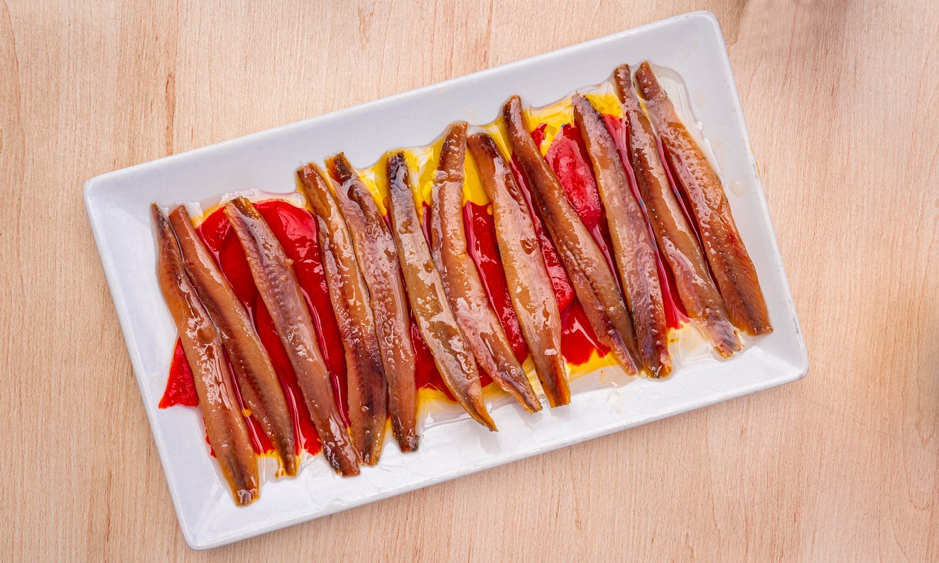 Anchovy fillets arranged neatly on top of red pepper slices, drizzled with olive oil, served on a rectangular white plate atop a light wooden surface—a beautiful presentation of one of those foods people learn to appreciate later in life.