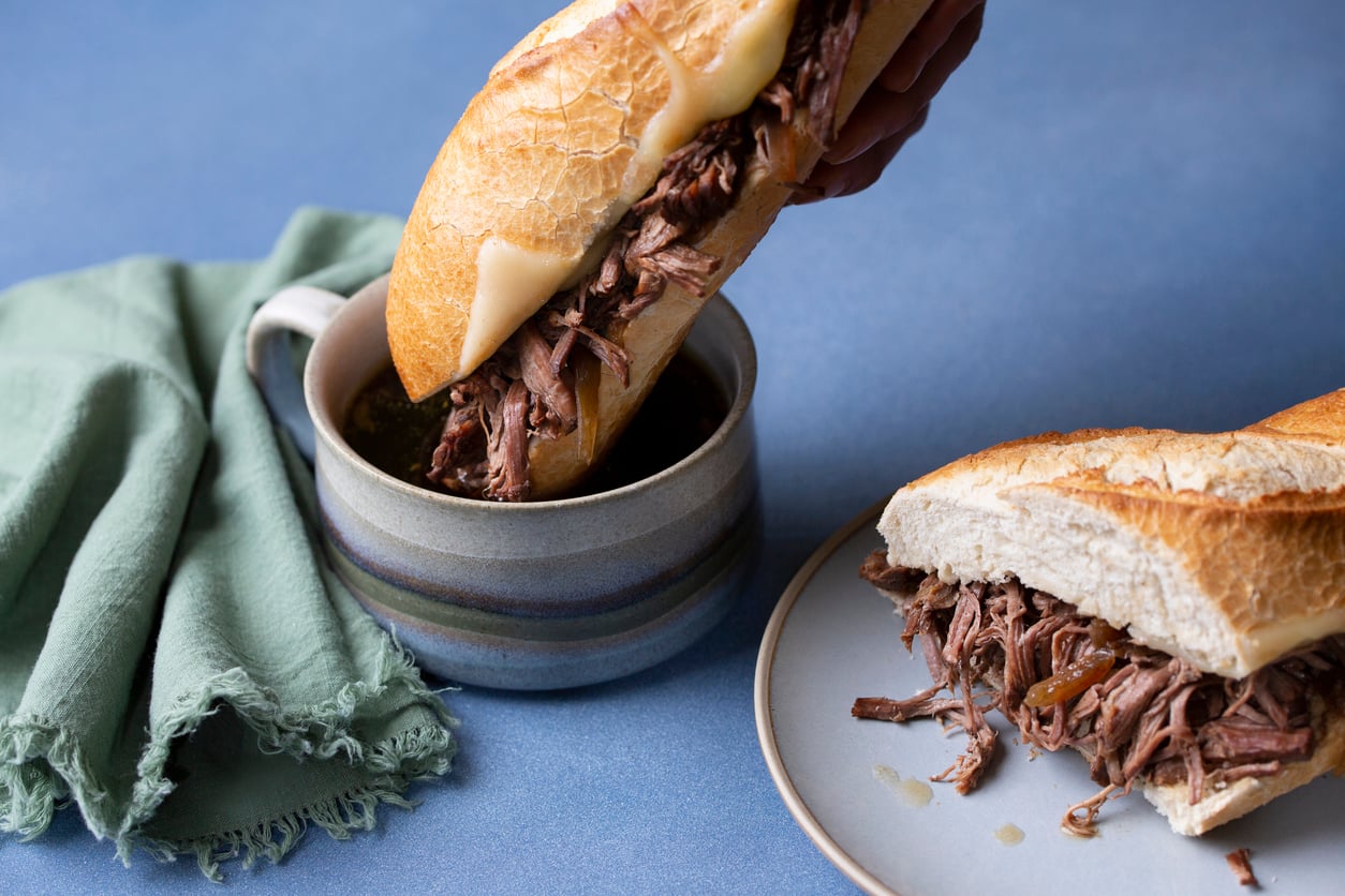 A hand dips a French dip sandwich filled with shredded beef into a cup of au jus. Another half sandwich sits on a plate nearby, with a green napkin on the table.