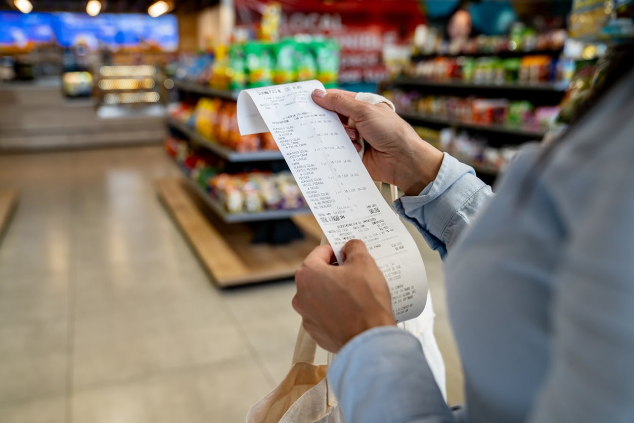 A person holds and reads a long receipt in a grocery store aisle, reflecting on their grocery habits, with shelves of snacks and various products visible in the background.