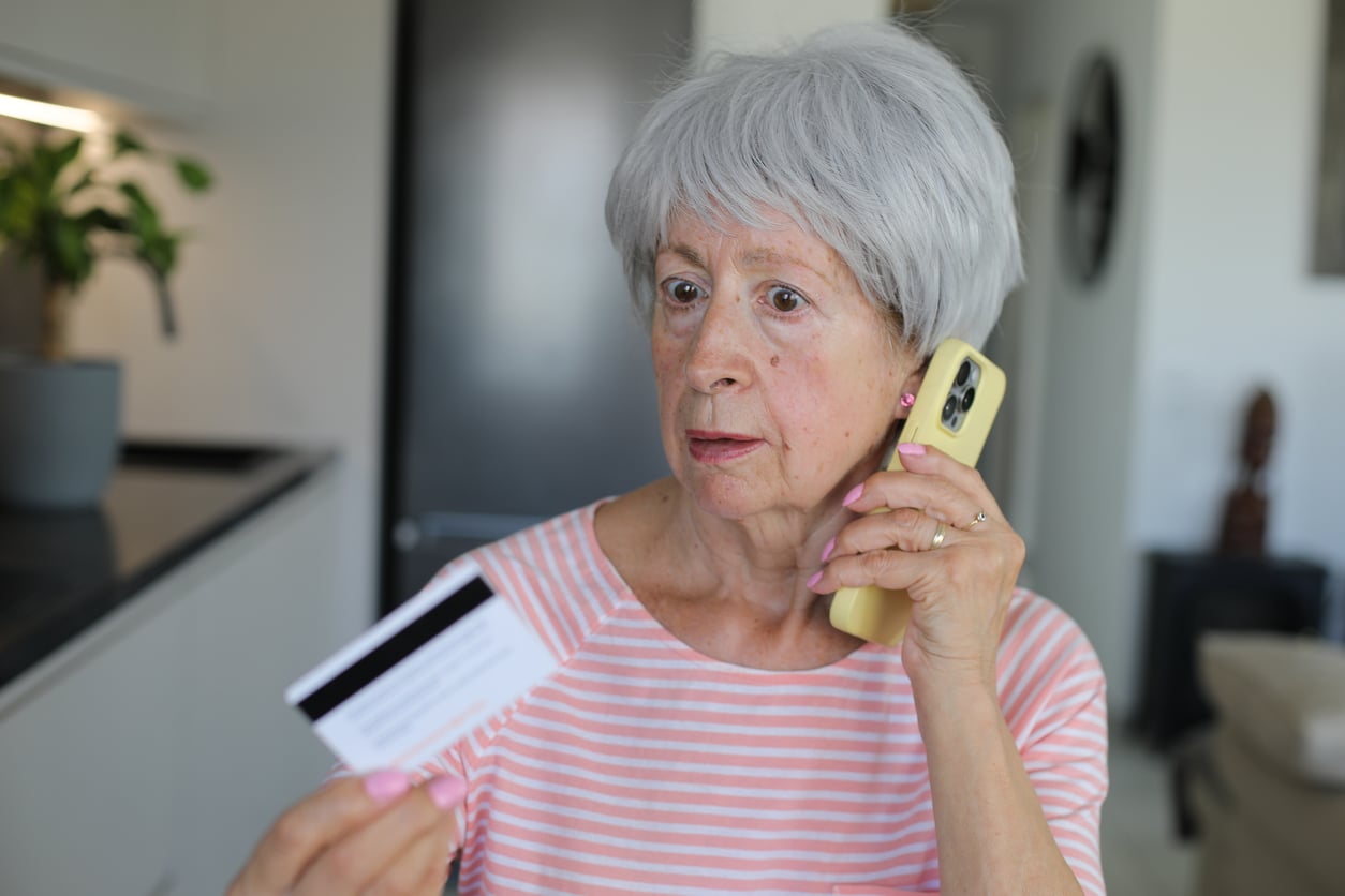 An older woman with gray hair holds a phone to her ear and looks at a credit card in her hand, appearing concerned or confused, perhaps contemplating her retirement money habits while standing in a home environment.