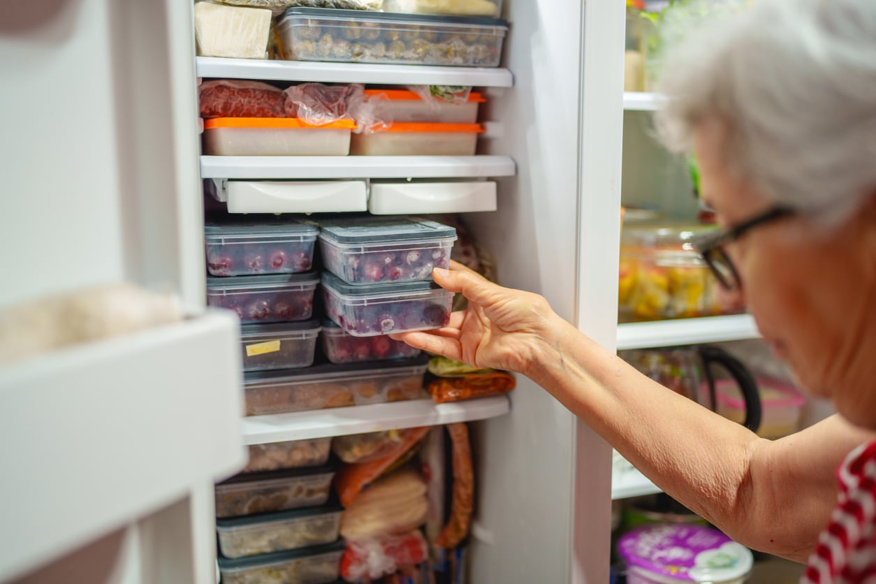 An older woman places a container of frozen food into a packed freezer filled with various foods in plastic containers and bags.