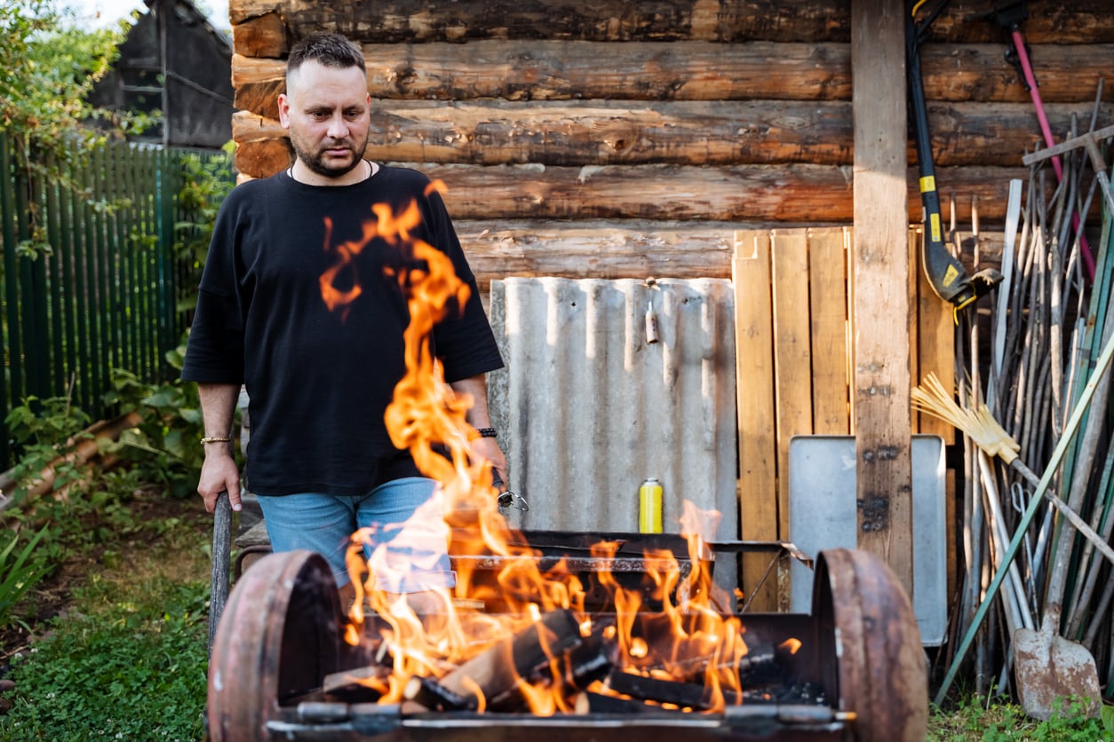 A man stands outdoors near a burning grill with flames rising, perhaps contemplating which foods to order to complement his barbecue. He is next to a rustic wooden shed, and garden tools are leaning against the wall behind him.
