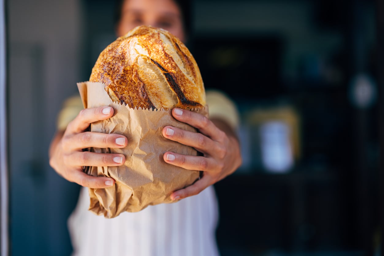 A person holds out a fresh loaf of bread wrapped in a brown paper bag, with their face blurred in the background.