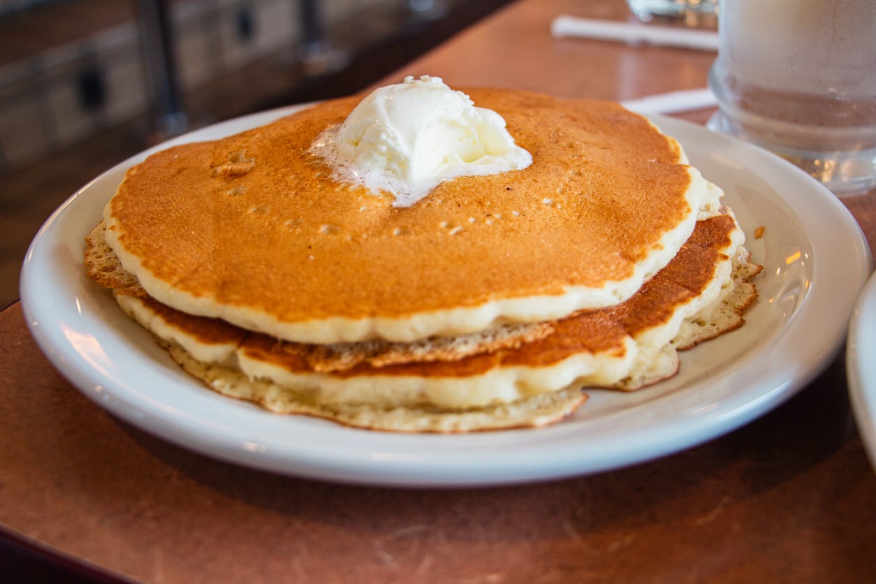 A stack of three golden-brown pancakes topped with a dollop of butter sits on a white plate on a brown table.