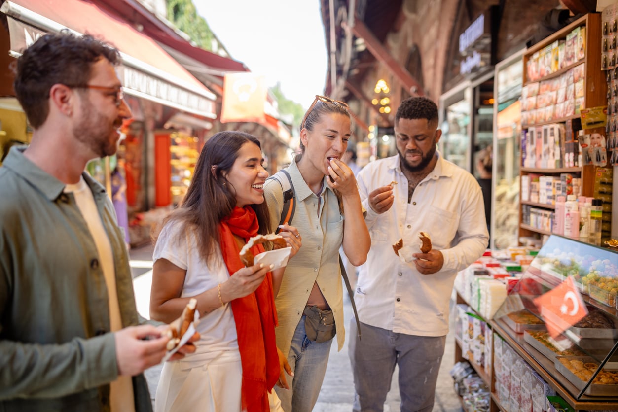 Four people stand together at an outdoor market, smiling and tasting local street food. They appear to be enjoying themselves, surrounded by colorful market stalls and various treats on display.