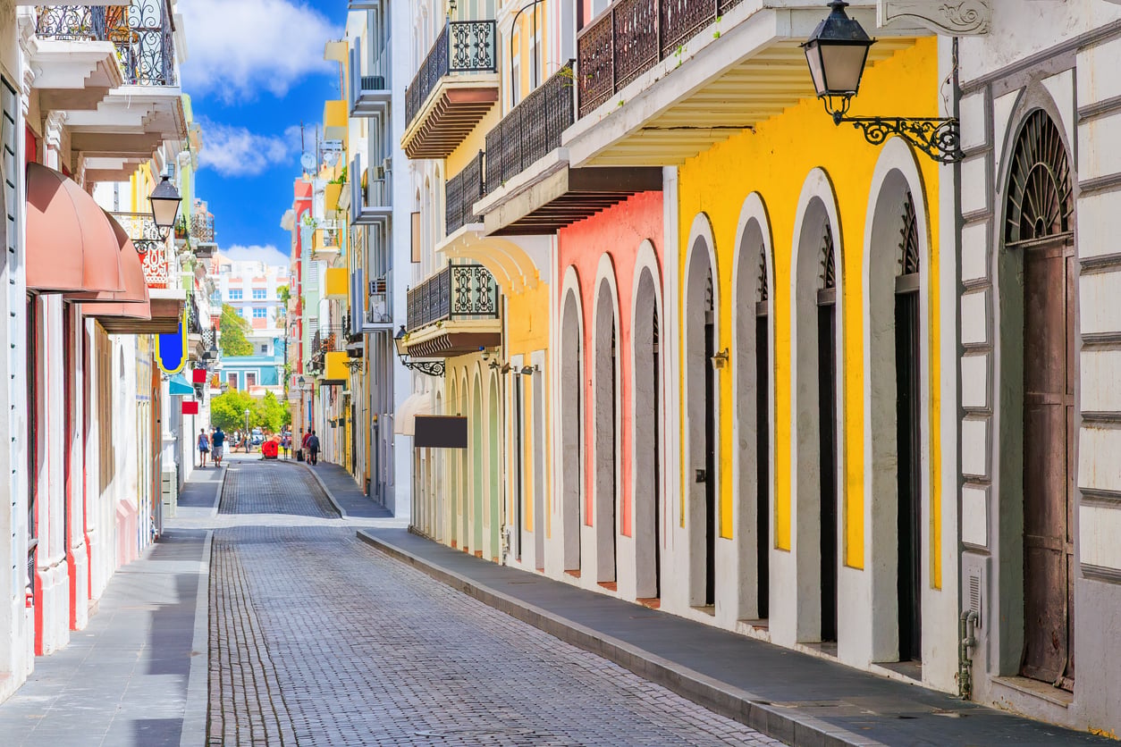 A narrow cobblestone street lined with colorful colonial buildings featuring balconies, arched doorways, and wrought iron details under a blue sky with white clouds.