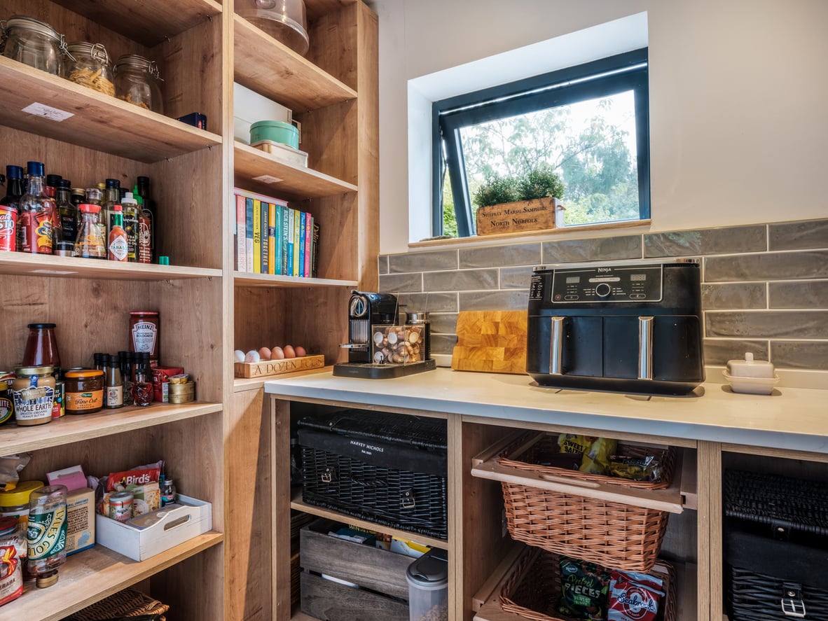A well-organized pantry reflecting mindful grocery habits features wooden shelves holding jars, spices, books, eggs, and baskets. The countertop displays an air fryer, coffee machine, a cutting board, and a small window with a potted plant.