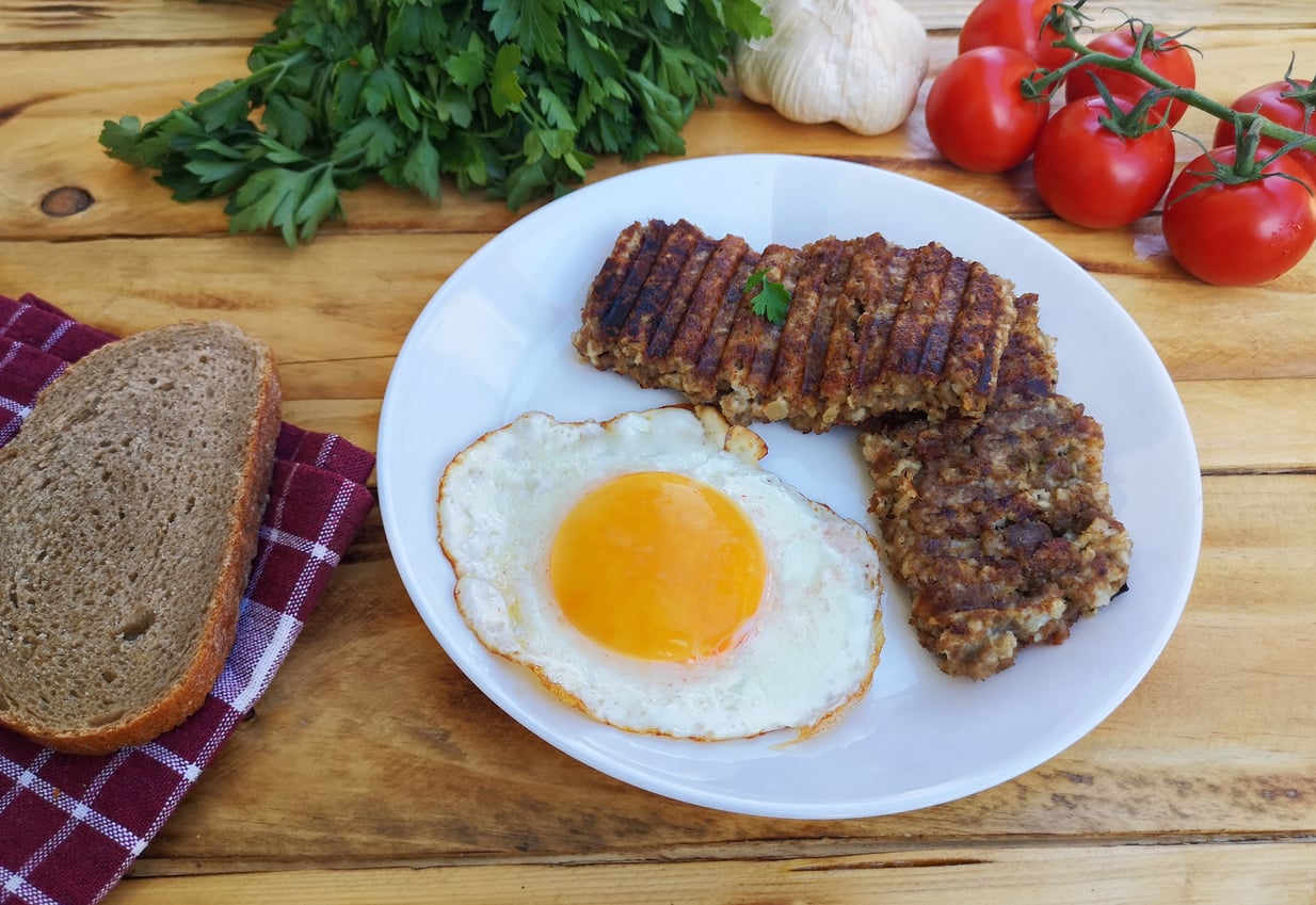 A plate with a sunny-side-up egg, grilled meat slices, parsley, tomatoes, garlic, a slice of brown bread, and a checkered napkin on a wooden table—an inviting showcase of restaurant specialties.