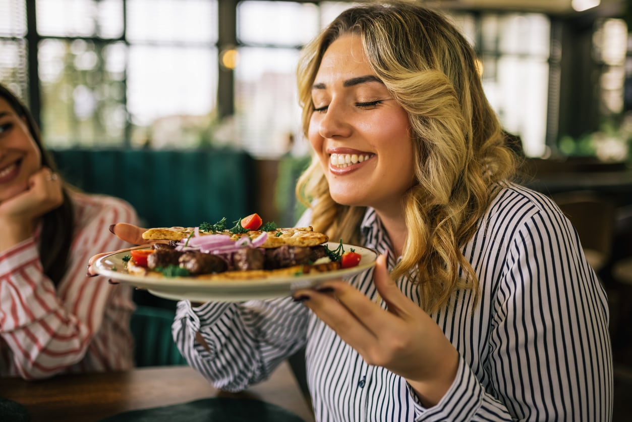 A woman with blonde hair and a striped shirt smiles while holding a plate of food close to her face, appearing to enjoy its aroma in a bright, cozy restaurant.