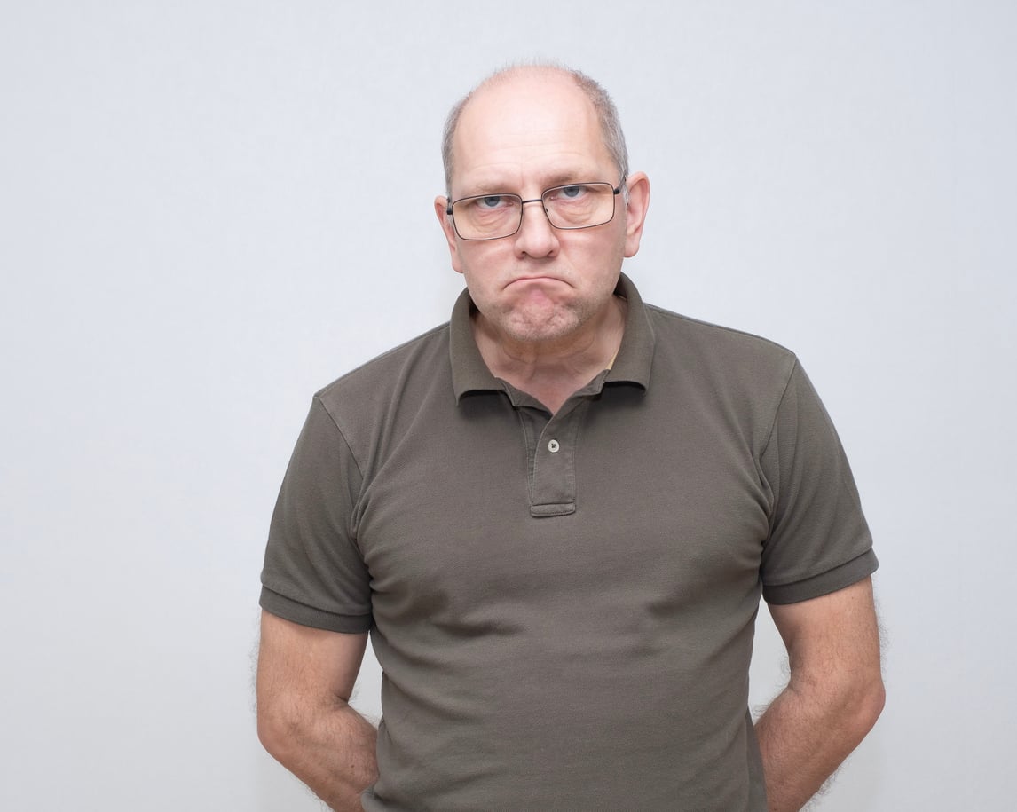A middle-aged man wearing glasses and a brown polo shirt stands against a plain light background, frowning with a displeased or unhappy expression.