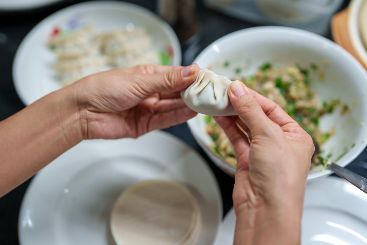 Hands are folding a dumpling wrapper around a filling. Bowls of filling, finished dumplings, and dumpling wrappers are visible on the table, illustrating the process of making homemade dumplings—one of the most beloved foods to order or prepare from scratch.