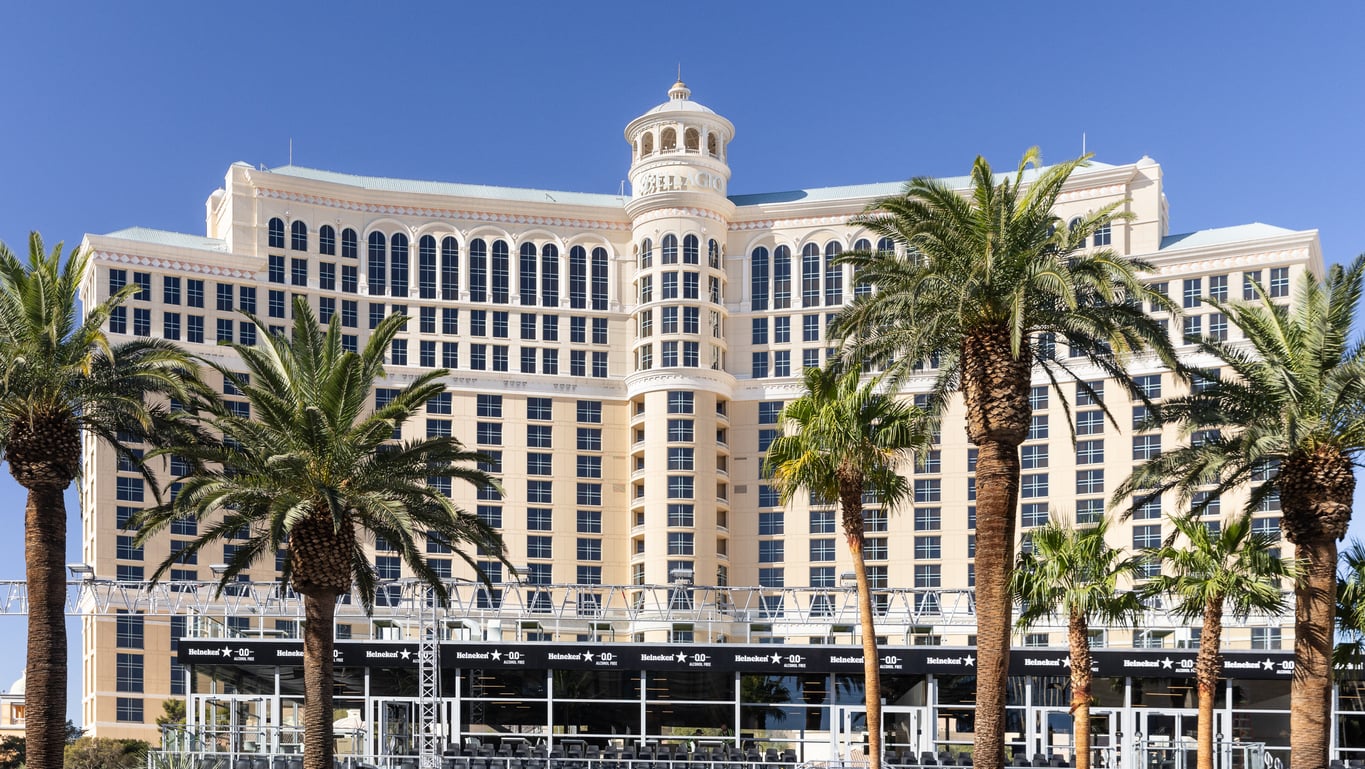 A large beige hotel building with many windows, tall palm trees in front, and a clear blue sky in the background.