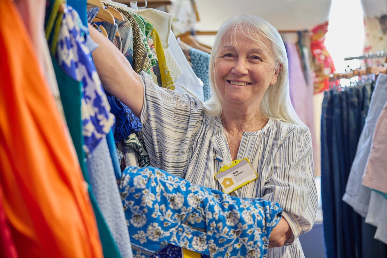 Smiling older woman with long white hair holds clothes on a rack in a brightly colored store. She wears a striped shirt and a yellow name tag, and appears to be arranging or sorting garments.