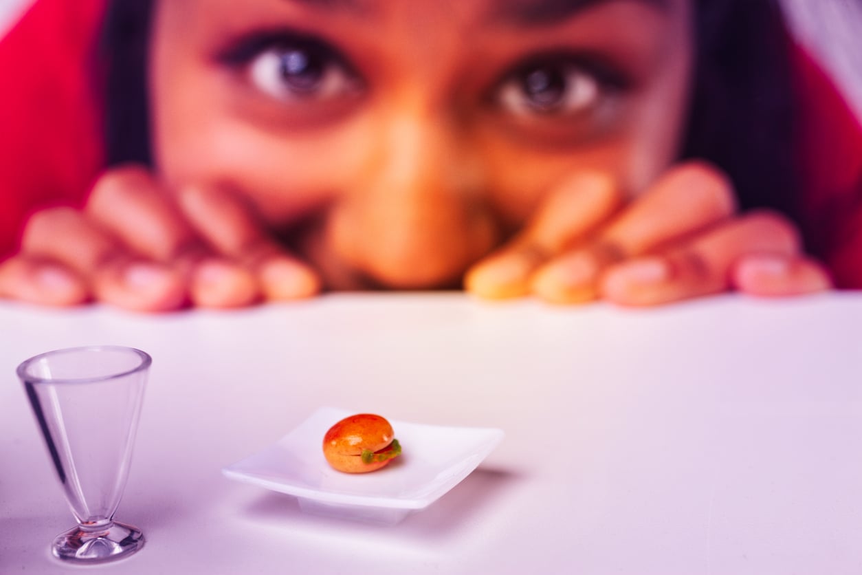 A person eagerly looks at a tiny hamburger on a small white plate, with a miniature glass nearby, all set on a white surface.