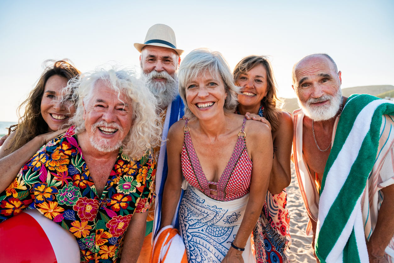 Six smiling older adults with towels and beachwear pose together on a sunny beach, enjoying a fun and joyful moment.