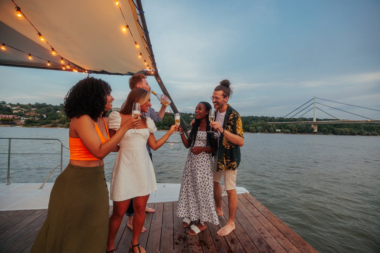 Five friends stand on a boat deck by the river, smiling and toasting drinks at sunset. Trees, a bridge, and string lights are visible in the background.