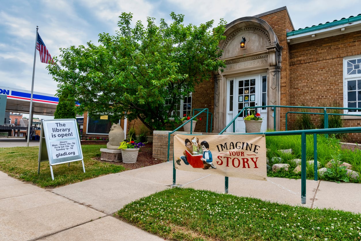 A brick library building with a tree, a sign reading “The library is open!” and a banner saying “Imagine Your Story” featuring children reading books, near a sidewalk and an American flag.