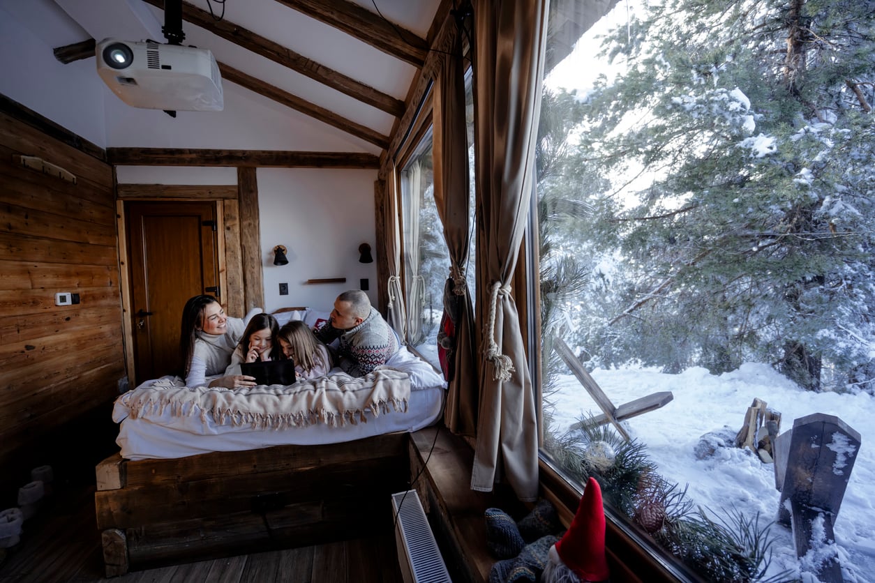 A family of four sits on a bed in a cozy wooden cabin, smiling and looking at a phone. Large windows show a snowy forest with pine trees outside. Warm sunlight fills the room.