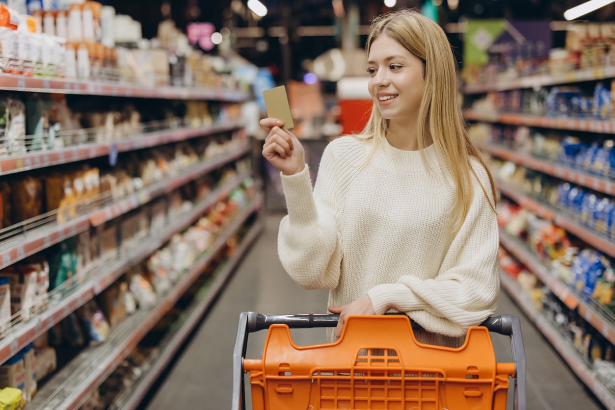 A woman in a white sweater holds a credit card while pushing a shopping cart down a grocery store aisle filled with products.