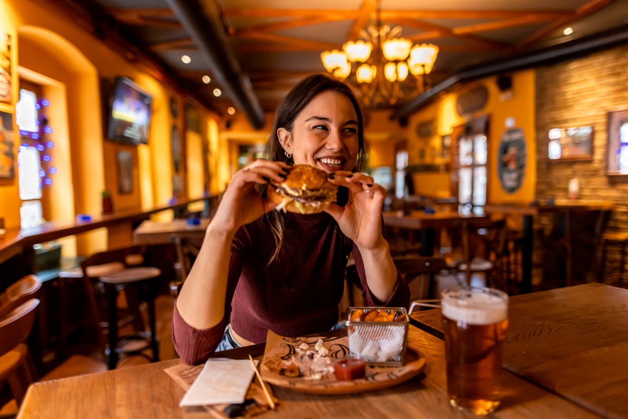 A smiling woman sits in a cozy restaurant holding a burger, with fries and a glass of beer on the table in front of her. The background features warm lighting and wooden decor.