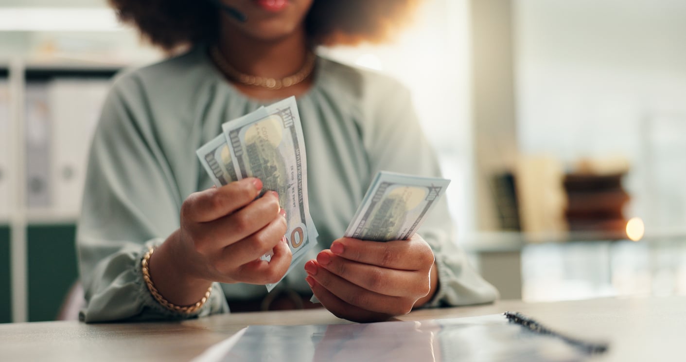 A person sits at a desk counting several $100 bills as a money-saving habits. Their face is partially out of frame, and they wear a light green blouse and gold jewelry. The background is softly blurred with office supplies visible.