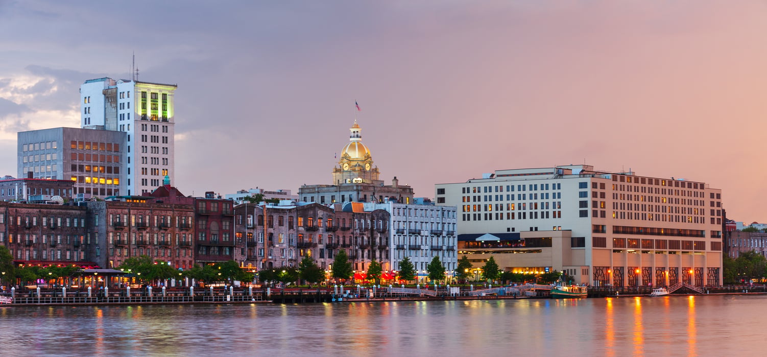 View of Savannah, Georgia’s historic riverfront at sunset, featuring old brick buildings, a domed courthouse, and lights reflecting on the water under a colorful sky.