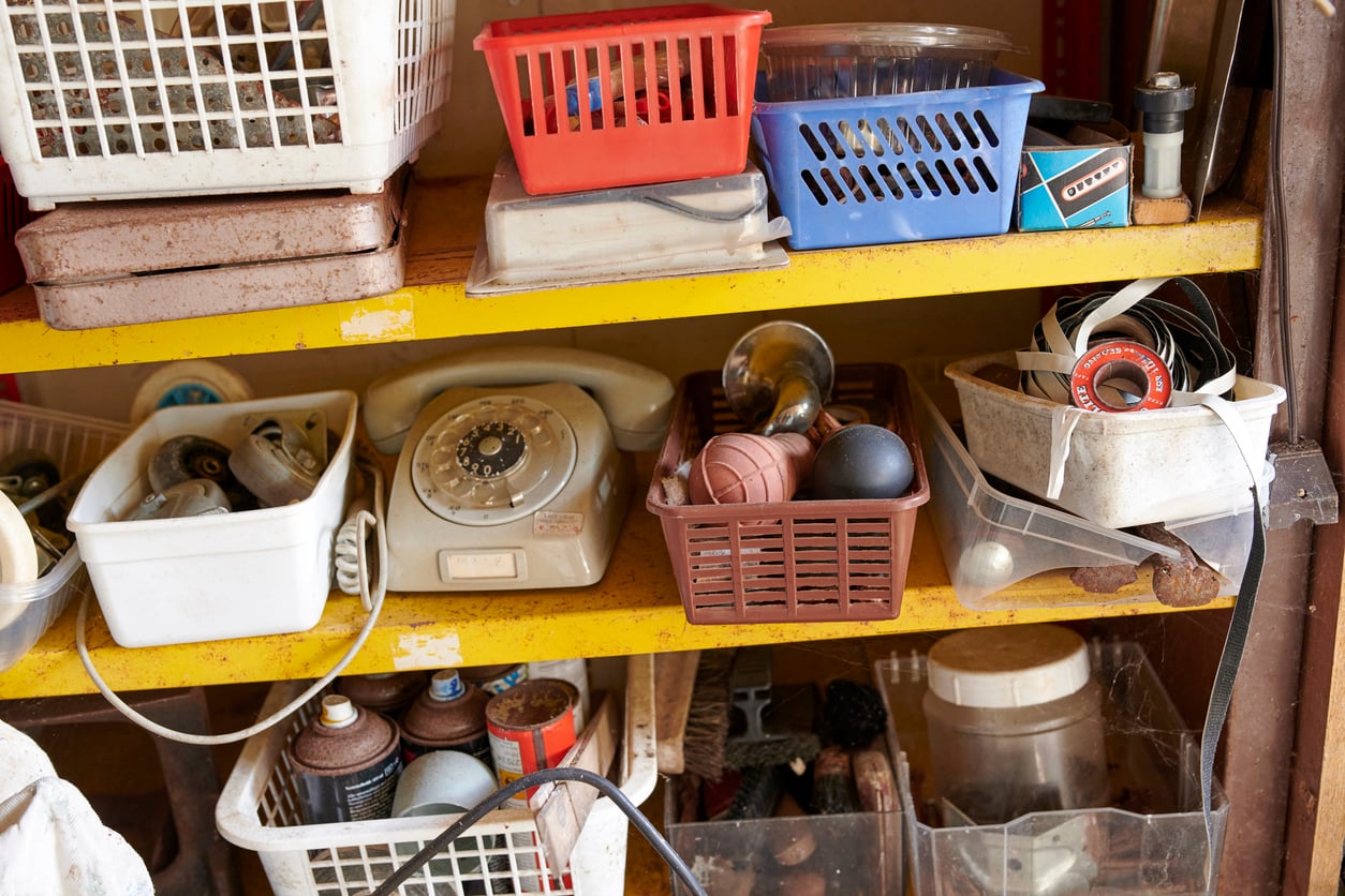 Yellow shelves with various bins holding an old rotary phone, spray cans, tools, light bulbs, wires, and other miscellaneous items in a cluttered workshop or storage area.