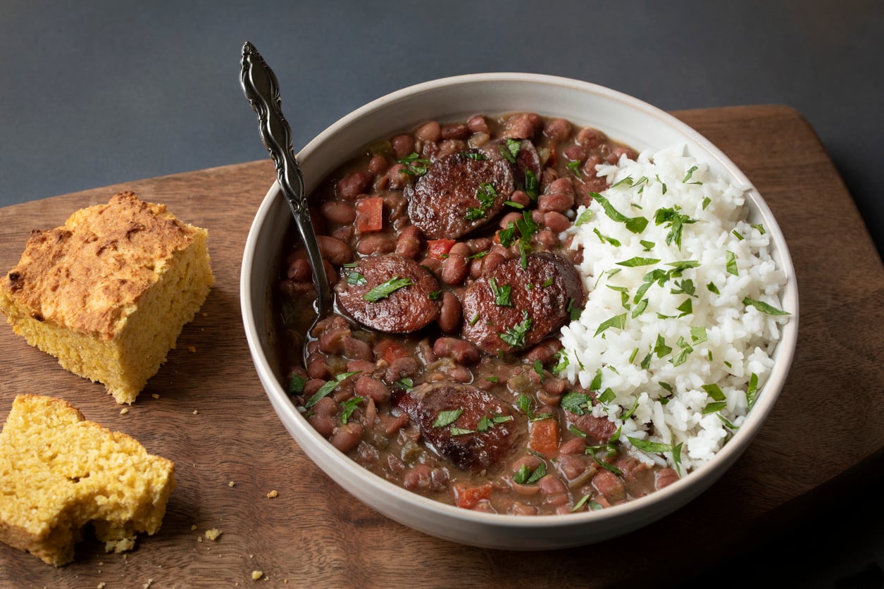 A bowl of red beans with sausage, garnished with parsley, served with white rice and a metal spoon. Pieces of cornbread sit on a wooden board beside the bowl.
