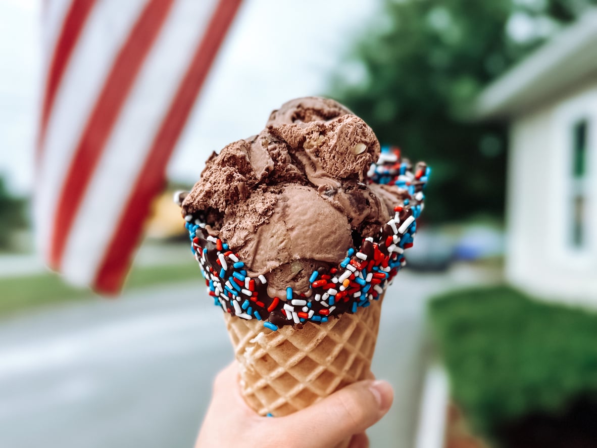 A hand holds a waffle cone with chocolate ice cream and red, white, and blue sprinkles—an irresistible combination that once might have ranked among everyday foods that used to be considered fancy. An American flag and green trees are blurred in the background.