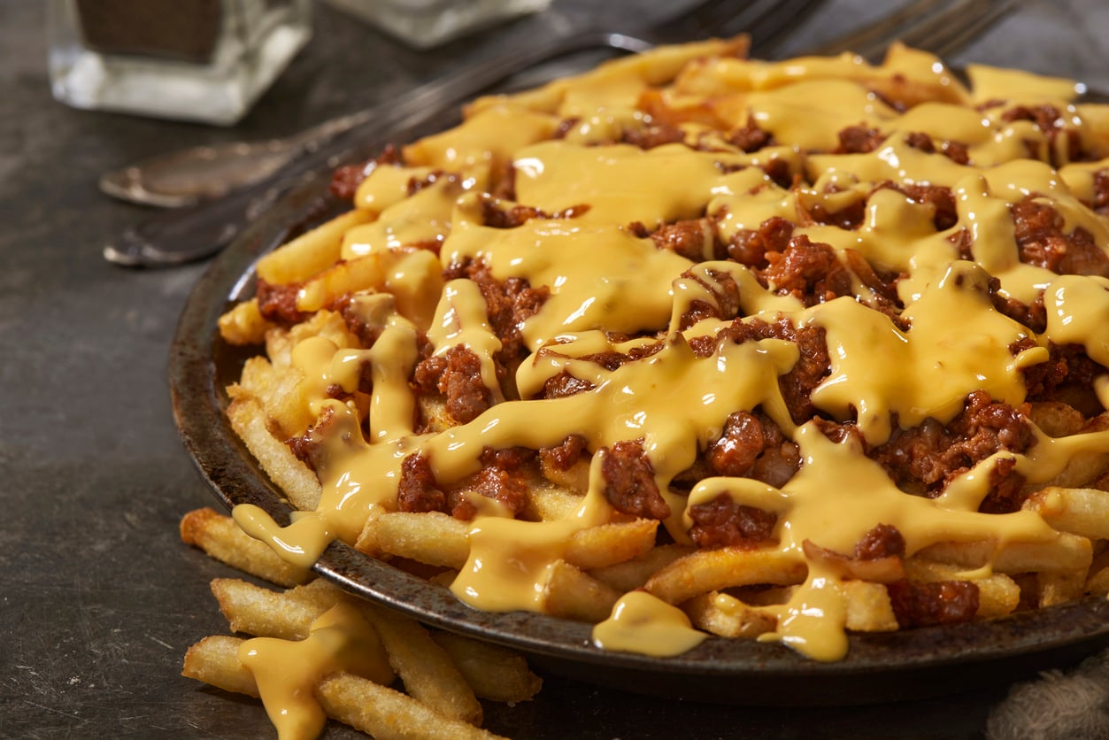 A plate of French fries topped with melted cheese and ground beef chili, served on a dark surface with utensils in the background.