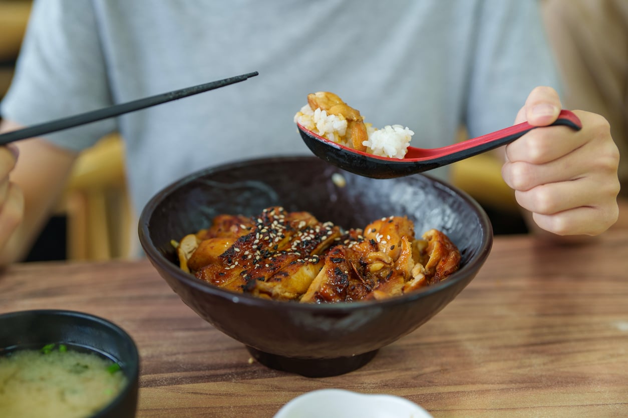 A person holding chopsticks and a spoon, eating rice and grilled chicken topped with sesame seeds from a black bowl, with a bowl of soup on the side.