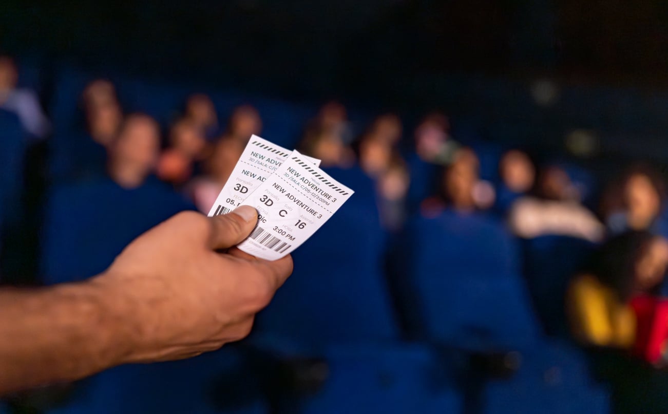 A hand holds two movie tickets in a theater, with people seated in blue chairs in the background, slightly out of focus.