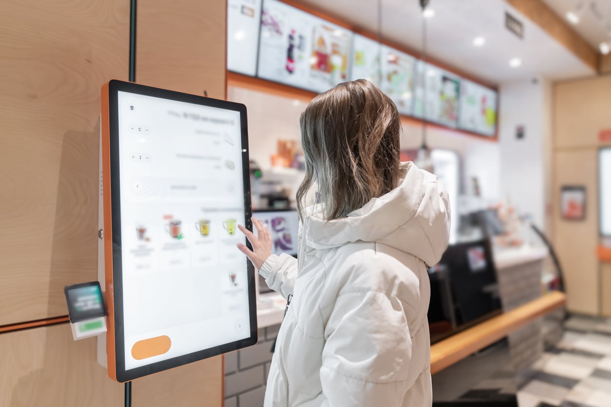 Person in a white jacket using a touchscreen self-service kiosk in a fast-food restaurant, with menu boards visible in the background.