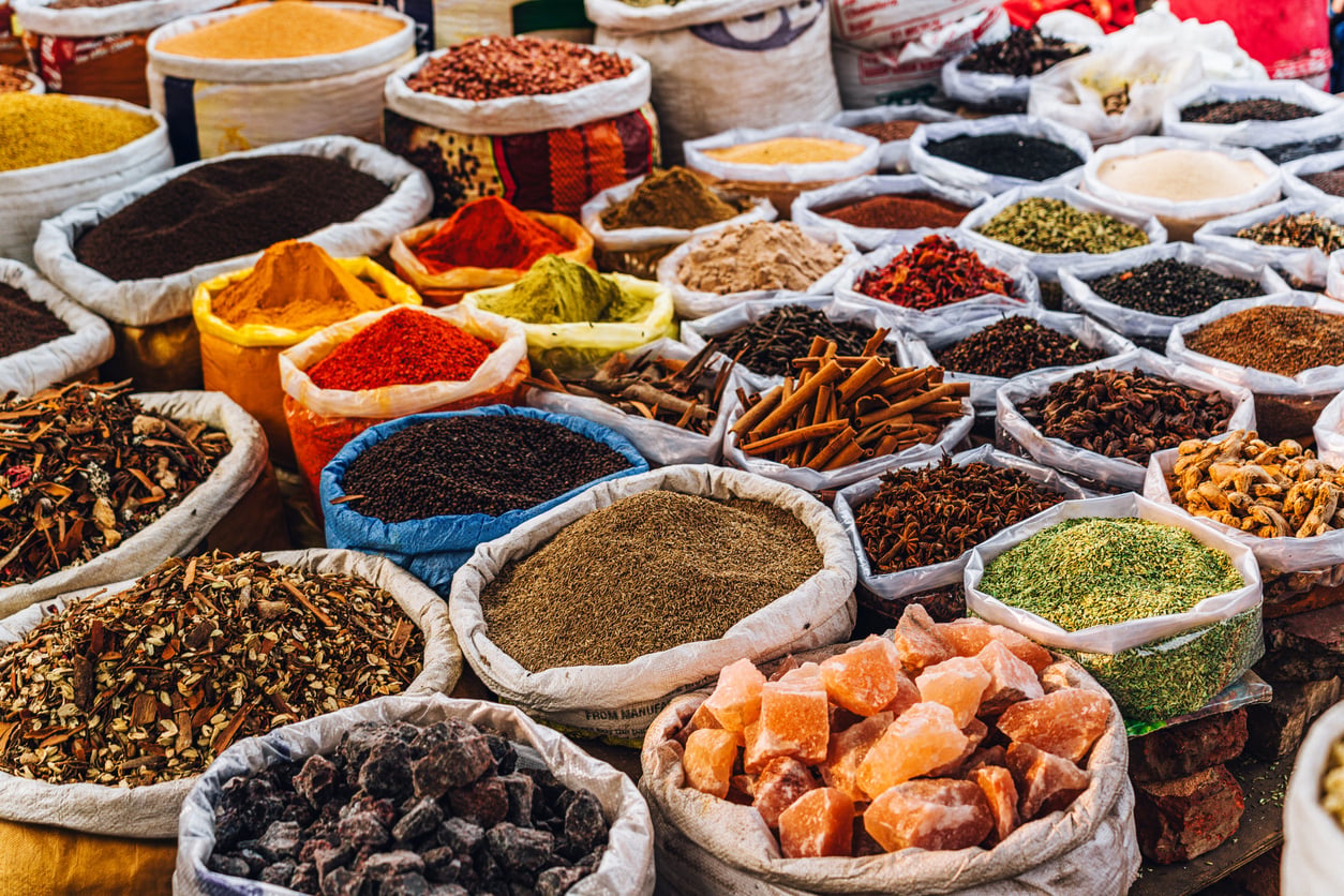 Colorful sacks filled with various spices and herbs are displayed at an outdoor market. The bags contain items like cinnamon sticks, dried herbs, seeds, and chunks of rock sugar, creating a vibrant, textured scene.