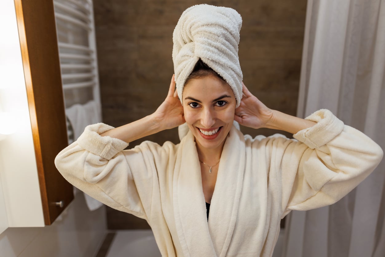 A smiling woman in a bathrobe stands in a bathroom with a white towel wrapped around her hair, holding the towel on both sides.