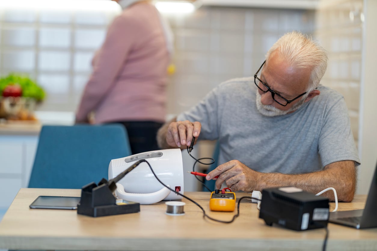 An older man wearing glasses repairs an electronic device at a table with tools; a woman stands in the background in a kitchen.