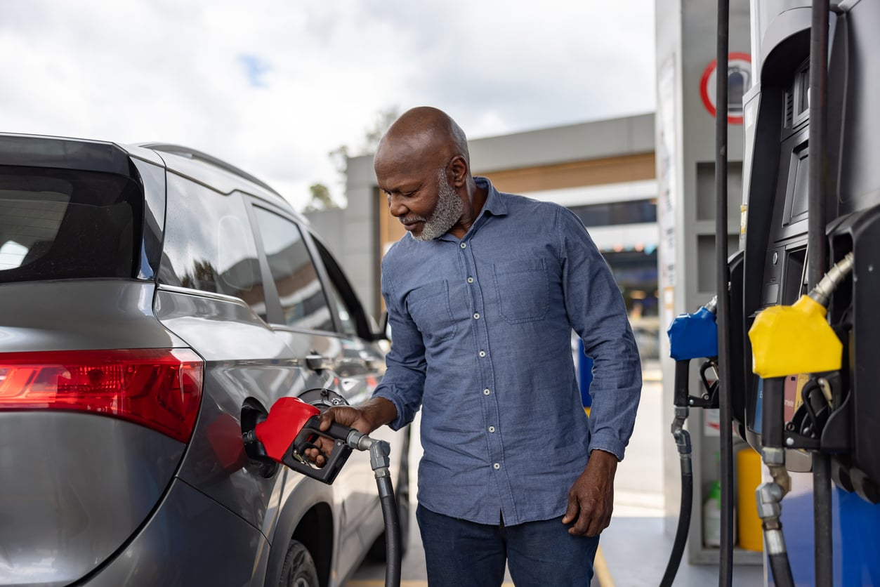 A man in a blue shirt is smiling while pumping gas into a gray SUV at a gas station. Fuel pumps and a canopy are visible in the background.