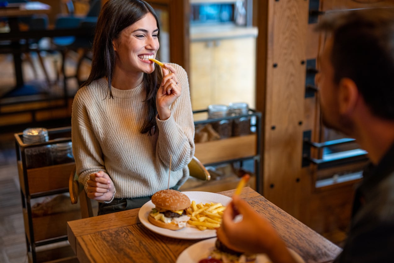 A woman smiles while eating a French fry across from a man at a restaurant table. Both have plates of burgers and fries in front of them. The setting appears casual and friendly.