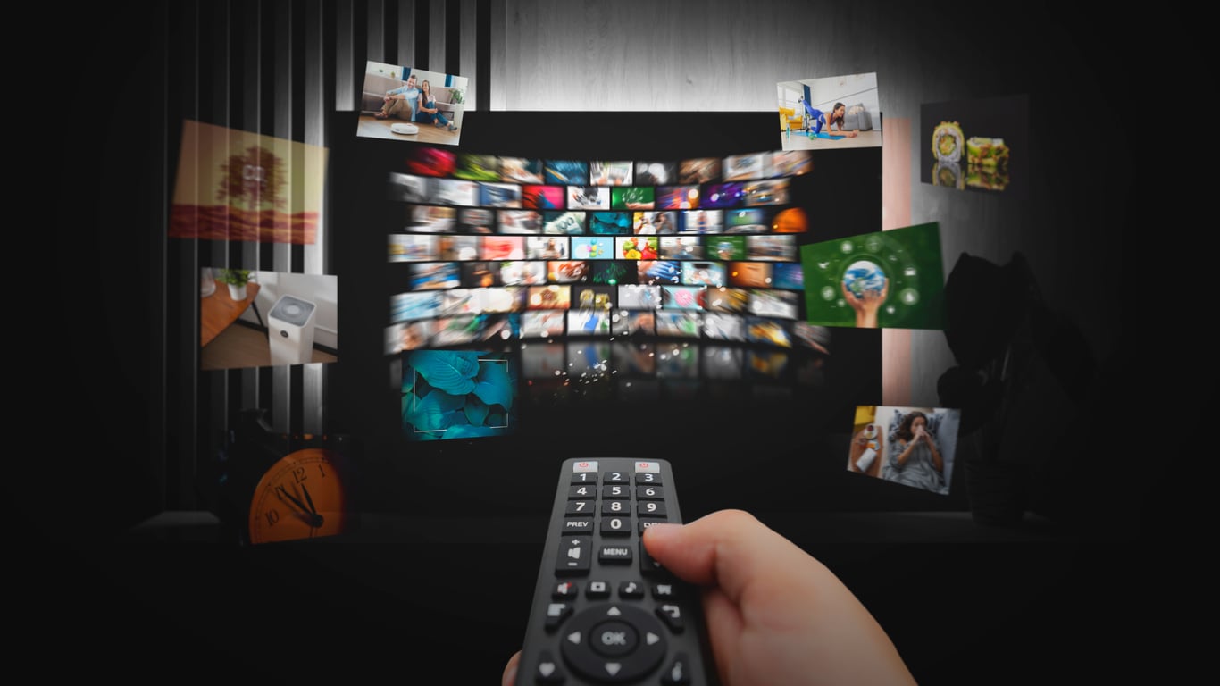 A hand holds a TV remote pointed at a screen displaying multiple streaming thumbnails, with various images like people, nature, and food floating around, suggesting a wide variety of digital content.