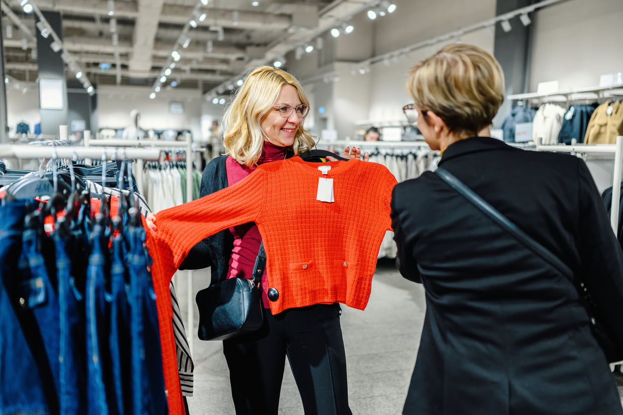 Two women shopping in a clothing store smile as one holds up a bright red sweater, discussing how clothing is often among the things families spend less on these days. Clothes hang on racks around them under bright store lighting.