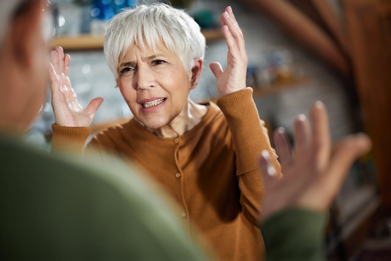 Older woman with short gray hair looks frustrated, raising her hands and frowning, while facing another person whose back is to the camera. They appear to be having a heated conversation indoors.